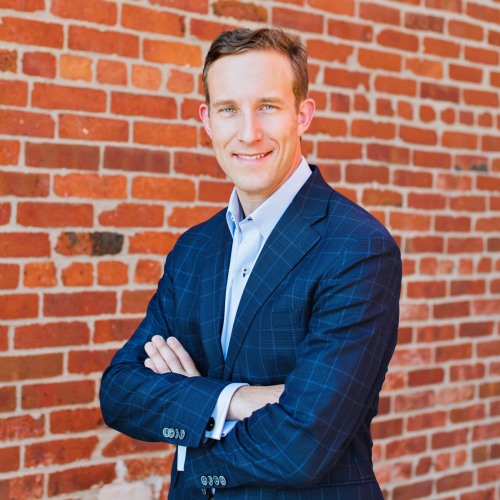 upper torso profile picture of an adult smiling, clean shaven with short dark hair, arms crossed in a blue casual suit standing in front of a red brick wall