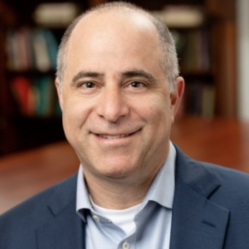 head and shoulders portrait of a smiling adult, clean-shaven, wearing a blue casual suit with bookshelves in the background