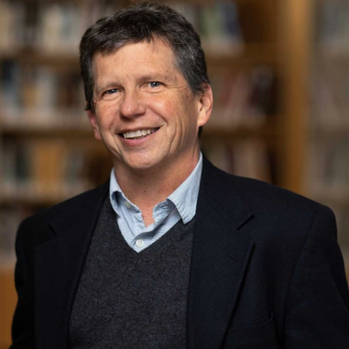 head and shoulders portrait of a clean shaved adult in business attire standing in front of bookshelves.
